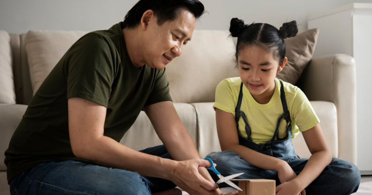 A father sitting on the floor with his daughter, helping her cut a cardboard box