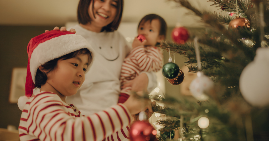 Closeup of a young boy hanging Christmas decoration on a tree, mother in the background with her hand on his shoulder while carrying a baby