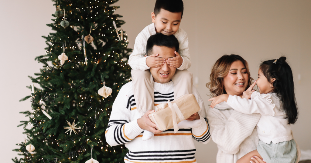 family of four in front of a Christmas tree, father with his son on his shoulders, mother carrying the daughter