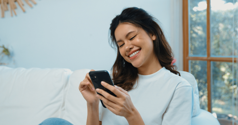 Send Money Internationally: Young woman sitting on the couch, smiling, holding and looking at her phone