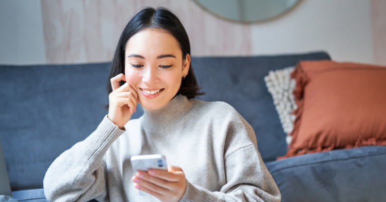 Young woman smiling, sitting down in front of sofa, looking at smartphone