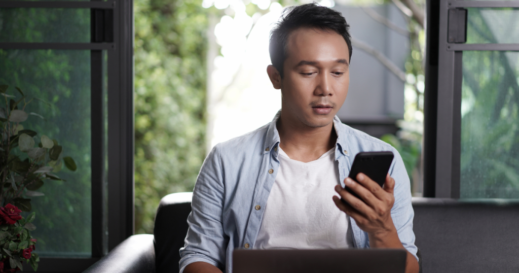 Man sitting on the couch in front of his laptop while looking at his phone in his hand