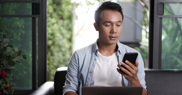 Man sitting on the couch in front of his laptop while looking at his phone in his hand