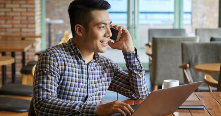 Man smiling while talking on the phone, sitting down in front of his laptop