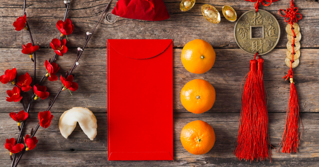 A wooden table with a red plant, a dumpling, a red envelope, tangerines, and Chinese coins tied with red string laying on it