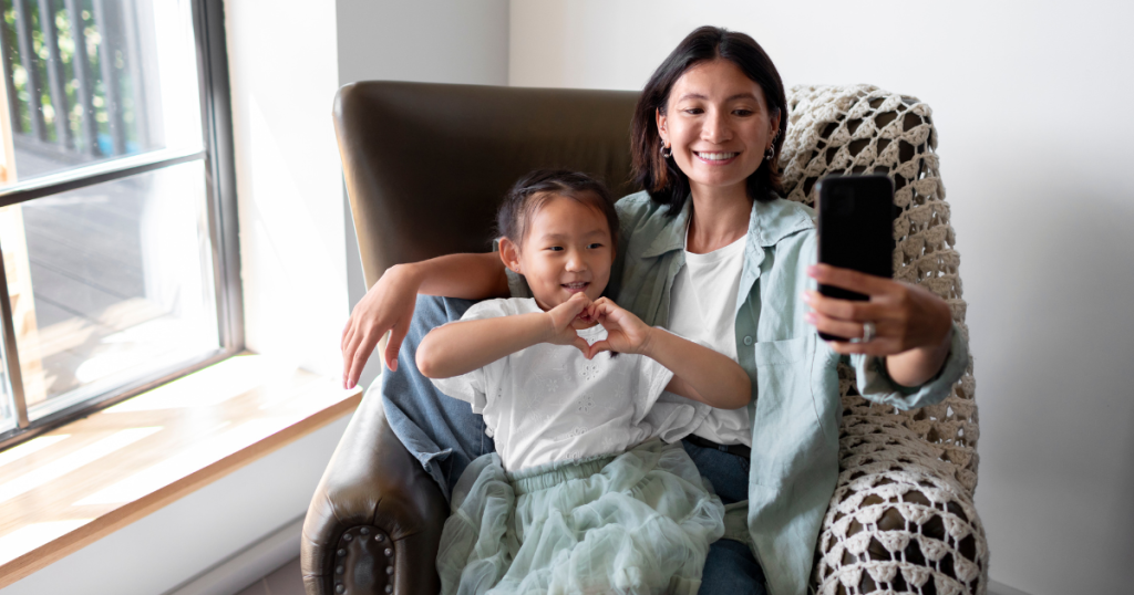 Mother and young daughter sitting on a couch together, mother's arm is outstretched in front of them both, holding a phone and smiling