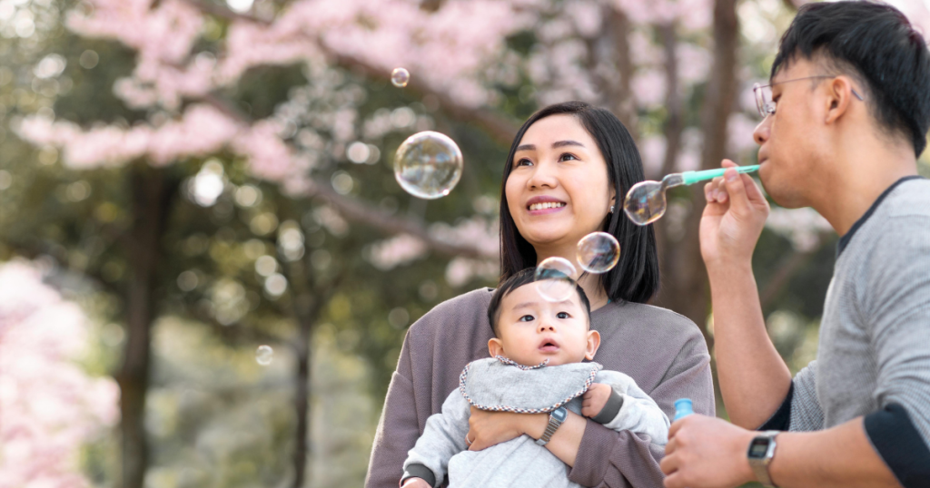 Family outdoors, man blowing bubbles while woman holds baby and stares at bubbles