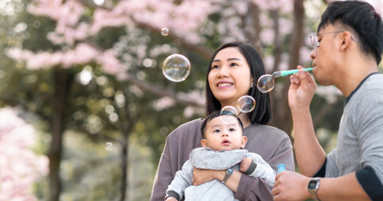 Family outdoors, man blowing bubbles while woman holds baby and stares at bubbles