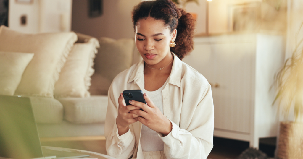 Woman scrolling on her phone while inside, sitting in front of her laptop