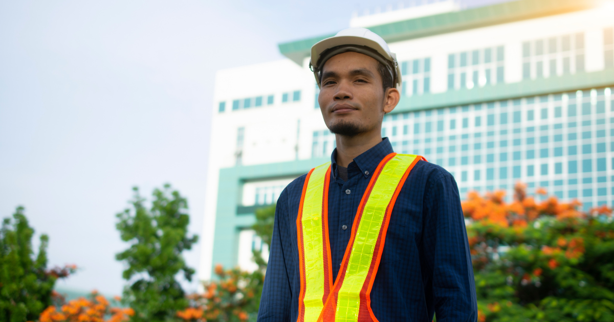 Man wearing safety vest and hardhat with building and trees in the background
